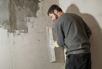 a plasterer man with a beard stands at a rough plastered wall with spatulas. 