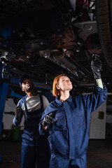 interracial workers of car service inspecting bottom of lifted auto in workshop.