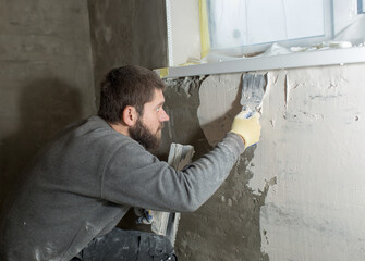 a man with a beard plasterer plasters a concrete wall under the windowsill with a spatula.