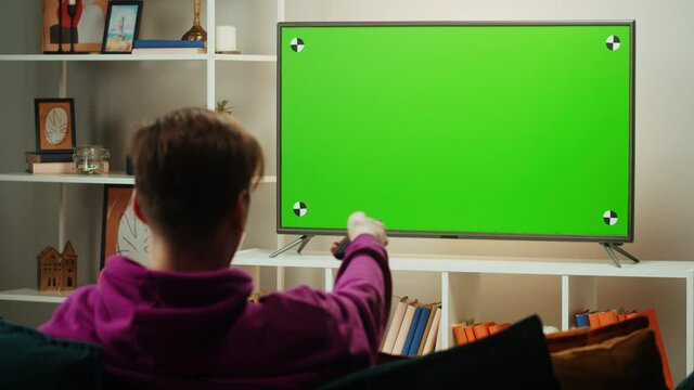 Man watching television in living room. Young guy sitting on sofa against tv with chroma green screen, back view. Male person switching channels using remote controller.