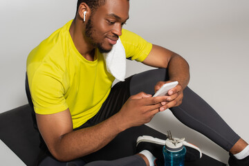 Fototapeta na wymiar pleased african american sportsman sitting on fitness mat and using smartphone near sports bottle isolated on grey.