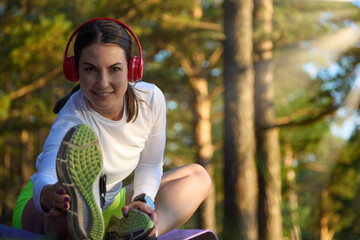 Young woman is warming up in the woods before jogging in red wireless headphones. Beautiful athletic girl doing fitness outdoors