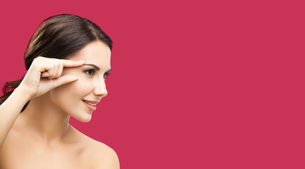 Fototapeta premium Skin care beauty concept. Portrait of smiling woman pointing brow or eye, touching skin, applying face cream, against red color background. Brunette model at studio shot.