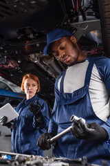 African american mechanic holding wrench near blurred colleague with digital tablet and car in garage.