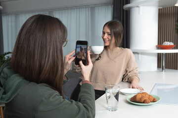 Two young women best friends sitting at cozy home eating croissants drinking coffee and taking photos with smartphone while they enjoying free time after long time they didnt see each other.