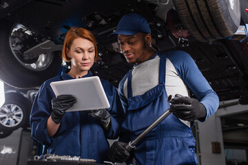 Multiethnic mechanics in gloves using digital tablet and holding wrench in car service.