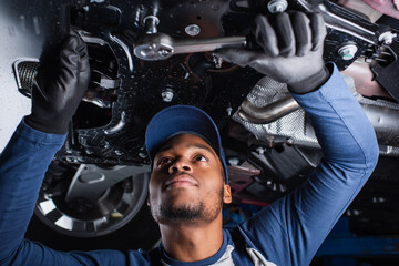 Young african american mechanic working with tool under car in garage.