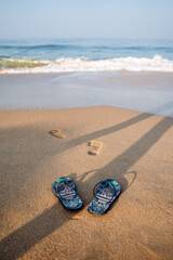 Fototapeta na wymiar Blue beach slippers on a sandy beach. Footprints of a man on the shore. Rest by the sea, summer adventures. Surf noise and sea foam