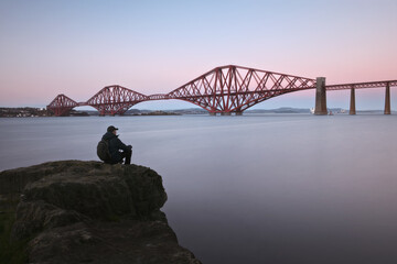 A man sits on a rock by the sea and looks at the sea and the big red old bridge. The Forth Rail...