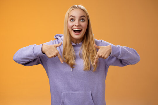 Excited fascinated european cute young girl blond hair smiling open mouth amused telling look down pointing downwards amazing cool new product recommend advertisement, orange background