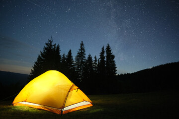 Bright illuminated tourist tent glowing on camping site in dark mountains under night sky with...