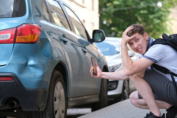Sad male driver photographing on sellphone camera dented vehicle on street side for insurance...