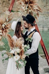 Bride and groom stand next to each other and smile, with a bouquet of flowers in boho style and...
