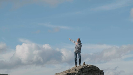 Funny man jumping on top cliff, celebrates climbing to mountain on sunny day. Beautiful background of blue sky, white clouds. Young enthusiastic man rejoices while dancing on top mountain