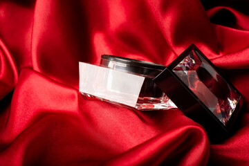 Cosmetic glass jar with a lid on red silk fabric background.