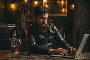 Fototapeta na wymiar Stylish rich man holding a glass of old whisky. Bearded man enjoying whiskey or cognac and doing his business at laptop.