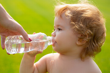 Baby kids drinking water from mother hands. Baby boy with curly blond hair drinking water in the park, holding plastic bottle, outdoor.