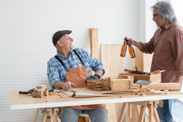 Two senior carpenter men Asian and Caucasian celebrating together with couple bottle of beer after finished their wood work at workshop. Working happily in retirement concept