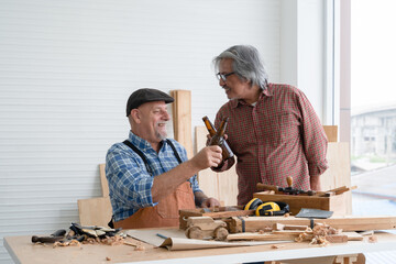 Two senior carpenter men Asian and Caucasian holding and cheers bottle of beer together after finished their wood work at workshop. Working happily in retirement concept