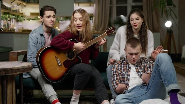 A young beautiful guy with a disability with two amputated stump hands in the company of friends and girlfriend is having fun with a guitar. Loft-style living room