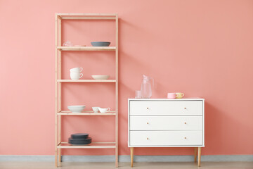 Fototapeta na wymiar Shelving unit and chest of drawers with dishes near pink wall