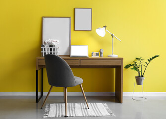 Modern workplace with laptop, frames and glowing lamp near yellow wall