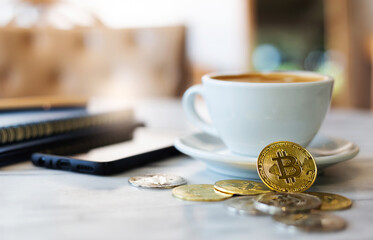 Select focus on .golden bitcoin in front of white coffee cup and blurred mobile phone and note book on desk. Concept trading on the cryptocurrency exchange.