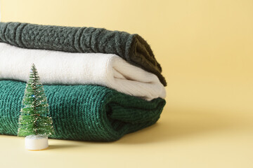 Stack of knitted sweaters and small Christmas tree on color background
