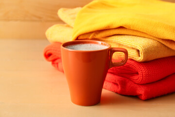 Stack of different cozy sweaters and cup of coffee on wooden background
