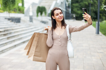 Cute smiling woman with shopping bags talking on makes selfie outdoors. Shopping concept