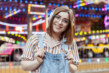 Obraz premium Cute smiling woman in denim overalls and glasses listens to music on headphones in an amusement park