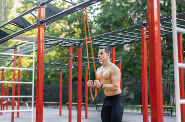 Obraz premium Athletic male athlete with a naked torso trains with a fitness elastic band on the workout site. Outdoor workout