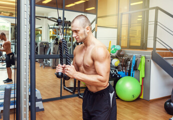 Muscular bodybuilder handsome man with naked torso trains triceps in a crossover cable exercise...