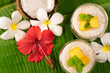 Tropical tapioca and coconut cream with mango dessert on green banana leaf background  frangipani and hibiscus flowers flat lay. © Inge