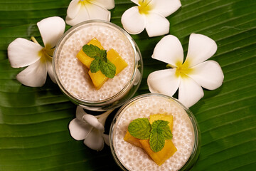 Fototapeta na wymiar Tropical tapioca and coconut cream with mango dessert on green banana leaf background with frangipani flowers flat lay top down view.