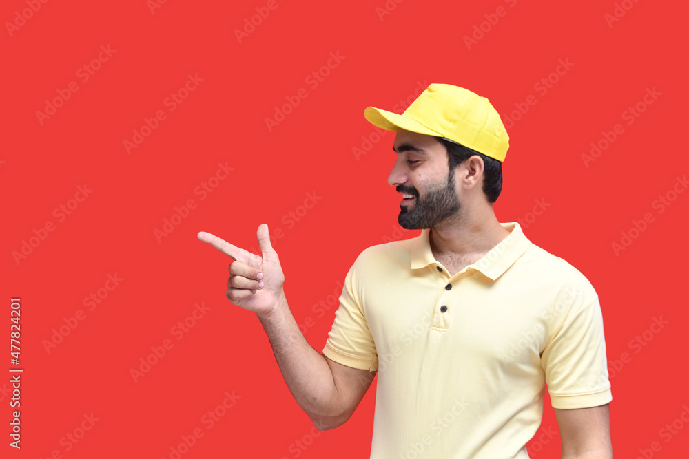 Wall mural young delivery man in yellow t shirt and cap smiling pointing aside up indian pakistani model