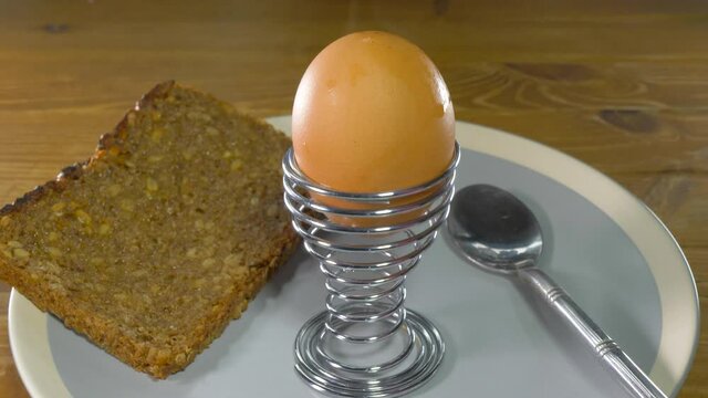 Closeup of a man’s hand placing a hot boiled egg in a spring cup / holder, onto a plate with a slice of toasted and buttered rye bread, next to a spoon.
