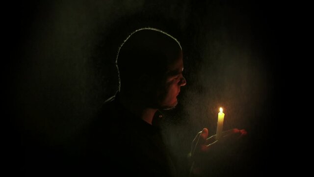 Man blowing out a candle in the dark. Falling water drops. Prayer with a candle. God's blessing. The man blows out the candle he is holding in front of him. Side view. 