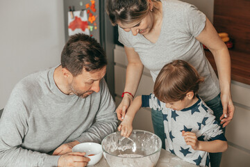 Cute little boy 4-5 years old and beautiful parents are happy while cooking cookies in kitchen at home. Mom, dad, son have baking party together,