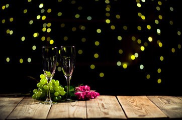 Preparations for celebrating Valentine's Day in romantic atmosphere with bokeh on the background. Still life made of two wine glasses standing on a wooden table and grape with roses lying next to them