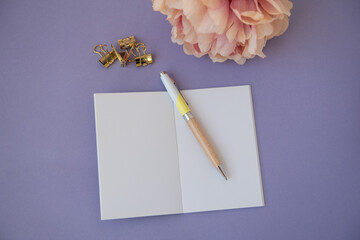Cahier ouvert sur une page blanche avec un stylo, une fleur rose et des pinces dorées sur un fond...
