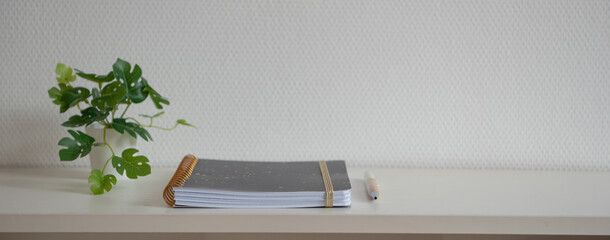 Cahier à spirale bleu et doré fermé et posé sur une étagère blanche avec un stylo devant un mur blanc avec une plante à feuilles verte