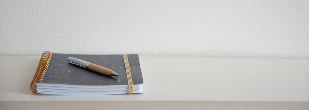 Cahier &agrave; spirale ferm&eacute; avec un stylo pos&eacute; dessus sur une &eacute;tag&egrave;re blanche et mur blanc - carnet simple et &eacute;pur&eacute;