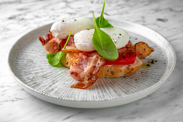 A healthy bruschetta breakfast with bacon, poached egg and fresh vegetables in a festive plate on a marble background. Restaurant banquet menu.