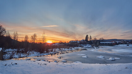 Winter evening, sunset over the river. Small dam, hydroelectric power station on an ice-covered river. Winter landscape