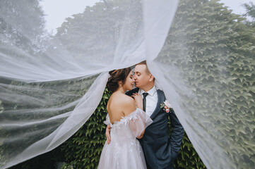 A young, stylish groom and a beautiful brunette bride embrace under a long fabric veil. Wedding portrait of lovers, smiling newlyweds. Photography, concept.