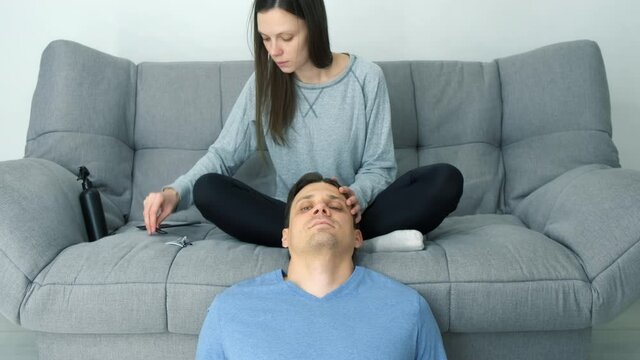 Woman is doing a man's hair while sitting at home on the couch. She combs his hair and pins it with hairpins preparing for braiding pigtails. Evening of a young married couple together.