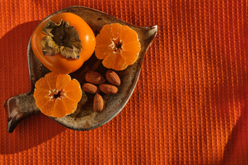 still life of tropical fruits: persimmons and tangerine cut in half and almonds on a clay saucer. Orange napkin background with space for text or logo, healthy food concept