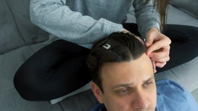 Woman is doing a man's hair while sitting at home on the couch. She sprays his hair with a styler and pins it with hairpins preparing for braiding pigtails. Evening of a young married couple together.