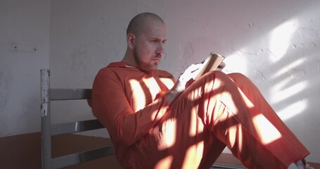 prisoner in orange overalls reading a book in a prison cell sitting on the bed.